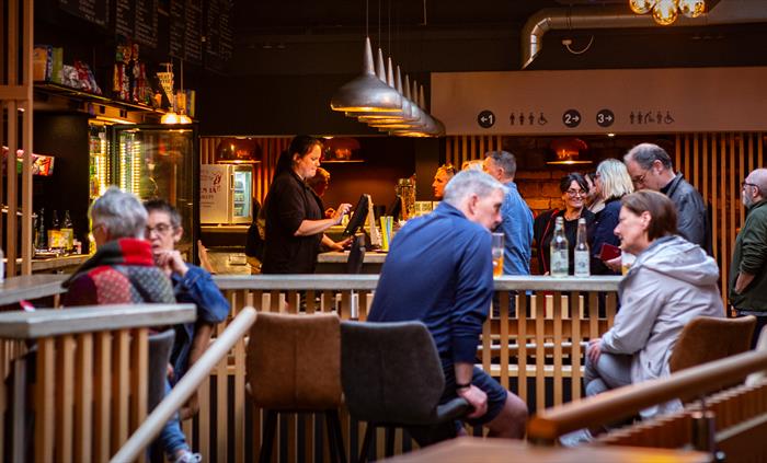 In the foreground, two pairs of people sit on high stools around a bar made of slated wood. In the background some people stand at a bar being served